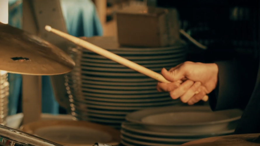 man holding drumsticks about the drum sitting in the middle of restaurant kitchen with stacks of plates in the background